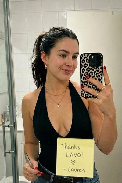 Woman takes selfie in bathroom, in background is a door, a toilet and a sink, on the mirror is a note saying 